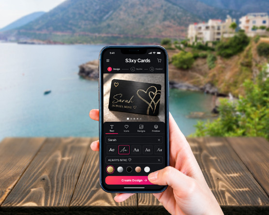 A hand holding a phone with the S3XY Cards designer open: a black card with a name and hearts in gold, and the text, icon, design and finish tools below it